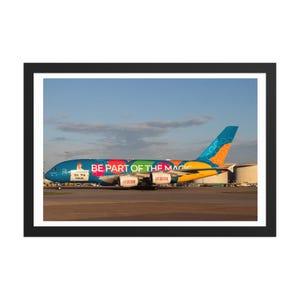 May include: A framed photograph of a large passenger aeroplane on the ground. The aircraft is painted in bright colours, including blue, orange, yellow, and green. The text "BE PART OF THE MAGIC" is visible on the side of the plane.