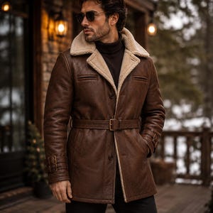 May include: A brown leather jacket with a cream-coloured shearling lining, a wide notched collar, and a matching leather belt at the waist. The jacket features long sleeves with buckled straps at the cuffs, two front pockets, and a button closure.