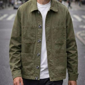 May include: An olive green button-up jacket featuring a classic collar, long sleeves with buttoned cuffs, and four large patch pockets on the front. The jacket is worn over a plain white t-shirt and paired with dark trousers for a layered look.