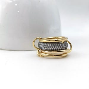 Op de afbeelding: Een stapelbare ringenset met een gouden bandontwerp en een centrale band bezet met kleine, sprankelende stenen. De ringenset wordt weergegeven op een wit oppervlak, met een wit keramisch object op de achtergrond.