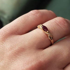 Peut inclure: Une bague en or ornée d'une pierre précieuse rouge foncé taille marquise au centre. L'anneau présente un design torsadé avec de petites pierres claires serties pavé sur les côtés, offrant un aspect délicat et complexe.