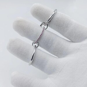 Pu&ograve; includere: Un anello color argento con una catena di pietre preziose rosa. L'anello &egrave; tenuto da una mano guantata. Il testo "RAWREETEXTILES" &egrave; visibile sull'anello.