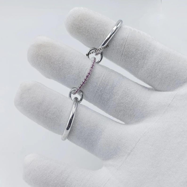 Pu&ograve; includere: Un anello color argento con una catena di pietre preziose rosa. L'anello &egrave; tenuto da una mano guantata. Il testo "RAWREETEXTILES" &egrave; visibile sull'anello.