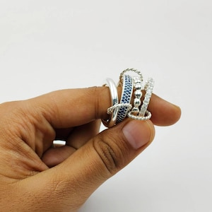 Puede incluir: Una colección de anillos de plata con varios diseños. Un anillo presenta una banda de pequeñas piedras azules, mientras que otro tiene un diseño de cuerda trenzada. Otros anillos incluyen una banda lisa y una banda con piedras transparentes.