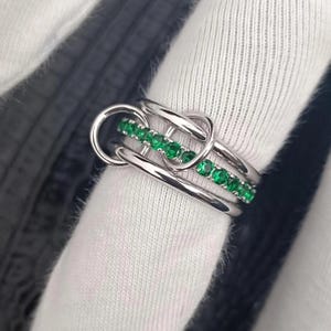 Op de afbeelding: Een zilveren ring met een uniek ontwerp. De ring heeft een rij kleine, heldergroene edelstenen tussen twee zilveren banden. De ring heeft een knoopachtig ontwerp met een cirkelvormig element.