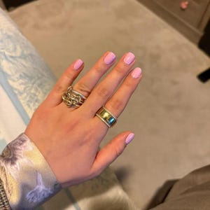 Peut inclure: Une main avec du vernis à ongles rose clair présente deux bagues. L'une est un design tissé en argent, et l'autre est une large bande dorée avec de petites pierres transparentes. La main est sur un fond neutre.