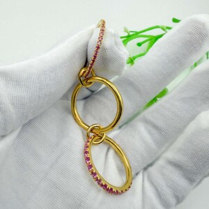 Op de afbeelding: Goudkleurige, verbonden oorbellen. Eén ring is ingelegd met kleine roze edelstenen. De oorbellen zijn verbonden door kleinere gouden ringen. De achtergrond is een wit oppervlak met een vleugje groen.