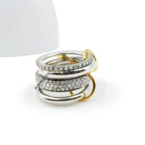 Op de afbeelding: Een meerbandige ring met zilver- en goudtinten. De ring heeft meerdere banden, waarvan sommige zijn bezet met kleine, heldere stenen. Het ontwerp bevat gouden accenten en een gelaagde uitstraling, wat een moderne en elegante look creëert.