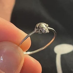Könnte beinhalten: Ein dünner silberfarbener Metallring mit einer kleinen, detaillierten Froschfigur, die oben am Ringband befestigt ist. Der Frosch ist mit sichtbaren Augen und Gliedmaßen geformt und nahtlos in das glatte, kreisförmige Ringband integriert.