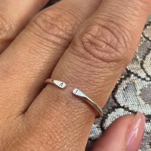 以下が含まれることがあります： 光沢のある仕上げのシルバー調のオープンリング。リングの両端はわずかに平らになっており、それぞれに文字が刻印されています。開口部の片側に「A」、もう片側に「J」の文字が配置されています。