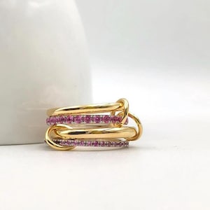 May include: A gold-colored ring set with three interlocking bands. Two bands are adorned with small, pink gemstones. The ring is displayed on a white surface, with a blurred white background.