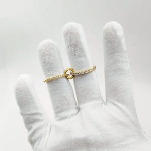 Op de afbeelding: Een goudkleurige ring met een cirkelvormig ontwerp en een band bezet met kleine, heldere stenen. De ring wordt getoond op een witte gehandschoende hand tegen een witte achtergrond. Het ontwerp van de ring bevat een klein, cirkelvormig accent.