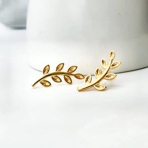 May include: A pair of gold-colored leaf-shaped earrings. Each earring features a delicate branch design with multiple leaves. The earrings are displayed on a white surface, with a blurred white background.