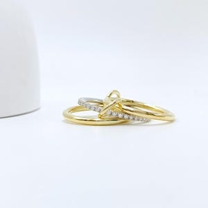 Op de afbeelding: Drie gouden ringen, waarvan één met een met diamanten bezette band, zijn op elkaar gestapeld. De ringen liggen op een wit oppervlak, met een wit object op de achtergrond. De ringen hebben een glanzende, gepolijste afwerking.