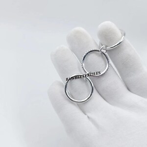 Pu&ograve; includere: Un anello color argento con tre cerchi interconnessi. Un cerchio &egrave; adornato con piccole pietre scintillanti. Il testo "BAWREETEXTILES" &egrave; visibile sull'anello. L'anello &egrave; presentato su sfondo bianco.