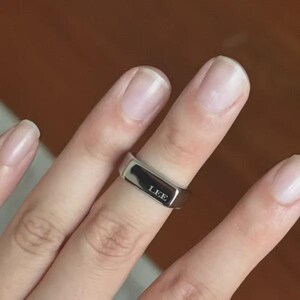Puede incluir: Un anillo de banda de metal plateado pulido con una cara rectangular plana. La palabra LEE está grabada en una fuente sans-serif limpia en la superficie frontal del anillo. La banda tiene un acabado liso y reflectante de diseño uniforme.