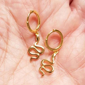 May include: A pair of gold-toned hoop earrings, each featuring a dangling snake-shaped charm. The earrings have a polished metallic finish and a hinged closure mechanism, showcasing a slender, curved serpent design that hangs from the circular hoop.