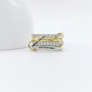 Op de afbeelding: Een stapel ringen met een mix van gouden en zilveren banden. Twee gouden ringen zijn versierd met kleine, heldere edelstenen. De ringen zijn in elkaar verweven, wat een uniek ontwerp creëert. De ringen liggen op een witte ondergrond.