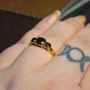 Op de afbeelding: Een gouden ring met drie rond geslepen zwarte stenen in een klauwzetting. De middelste steen is groter dan de twee zijstenen, allemaal gemonteerd op een gepolijste gouden band die de vinger omsluit met een gladde, metalen afwerking.