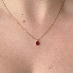 Könnte beinhalten: Eine zarte goldene Halskette mit einem einzelnen, kleinen, tropfenförmigen roten Edelsteinanhänger. Der Anhänger ist in einer Fassung aus goldfarbenem Metall befestigt und hängt an einer dünnen, polierten Gliederkette am Schlüsselbein.