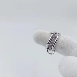 Pu&ograve; includere: Un anello d'argento con un design a doppia fascia, con una fila di piccole pietre rosa e un piccolo anello pendente. L'anello &egrave; esposto su sfondo bianco. Il testo "BAWREETEXTILES" &egrave; visibile sull'anello.