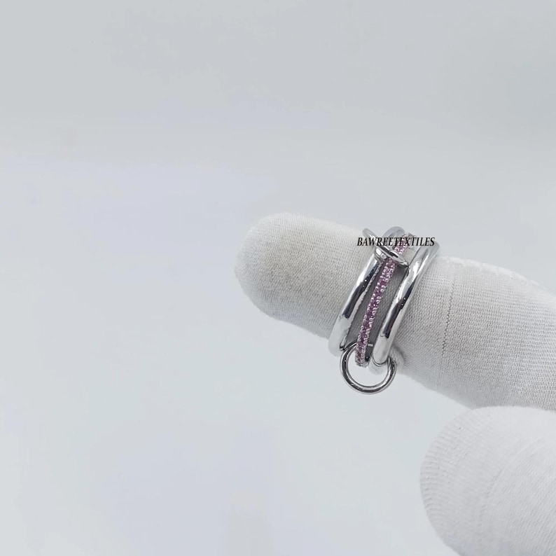 Pu&ograve; includere: Un anello d'argento con un design a doppia fascia, con una fila di piccole pietre rosa e un piccolo anello pendente. L'anello &egrave; esposto su sfondo bianco. Il testo "BAWREETEXTILES" &egrave; visibile sull'anello.