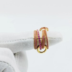 Op de afbeelding: Een verzameling goudkleurige ringen, elk versierd met kleine, levendige roze edelstenen. De ringen zijn gerangschikt in een gelaagd ontwerp, waardoor een visueel aantrekkelijk en ingewikkeld sieraad ontstaat. De edelstenen zijn dicht op elkaar gezet.