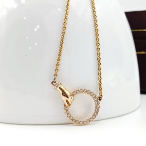 Può includere: Collana dorata con un ciondolo a forma di mano che tiene un cerchio ornato di piccoli cristalli trasparenti. La catena sottile e il ciondolo sono posti su uno sfondo bianco, evidenziando il design e la brillantezza dei cristalli.