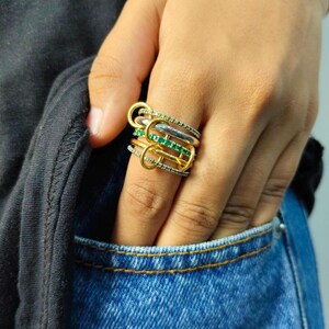 Op de afbeelding: Een meerlagige ringenset met gouden, zilveren en smaragdgroene accenten. De ring heeft een uniek ontwerp met meerdere banden en decoratieve elementen. De sieraden worden op de vinger van een persoon getoond, waarbij de ingewikkelde details en het kleurcontrast worden getoond.