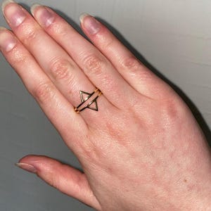 May include: A gold-toned ring featuring a geometric design composed of two open triangles stacked vertically. The triangles are connected by a thin band that encircles the finger, creating a symmetrical, open-frame appearance on the finger.