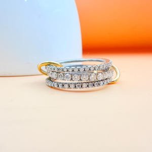 Op de afbeelding: Een zilveren ring met drie banden, elk bezet met kleine, heldere edelstenen. De banden zijn verbonden met goudkleurige connectoren. De ring staat tegen een perzikkleurige achtergrond met een witte en oranje achtergrond.