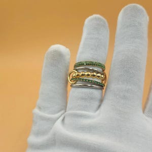 Op de afbeelding: Een stapelbare ringenset met meerdere banden in goud- en zilverkleuren. De ringen zijn versierd met kleine, sprankelende groene stenen en goudkleurige bollen. Gepresenteerd op een witte handschoen.