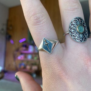 May include: A silver ring with a diamond-shaped face featuring a sword design on a dark blue background. Another ring with an openwork design and a central green stone is also visible.