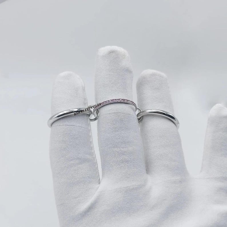 Pu&ograve; includere: Un set di anelli d'argento, uno con una delicata fascia di piccole pietre rosa, collegato a un secondo anello d'argento liscio da una barra curva. La barra &egrave; incisa con la scritta "BAUBLE TEXTILES".