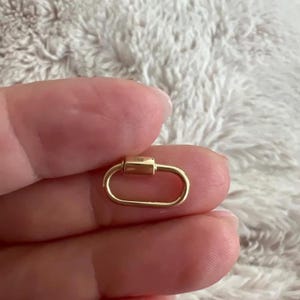 Puede incluir: Un cierre de mosquetón ovalado de color dorado. El cierre tiene una barra rectangular con un pequeño detalle cuadrado. El mosquetón está en la mano de una persona.