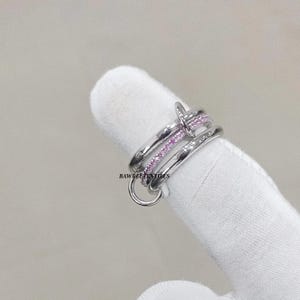 May include: A silver-colored ring with a nail-inspired design. The ring features three bands, one of which is adorned with small pink gemstones. The ring is shown on a white surface.