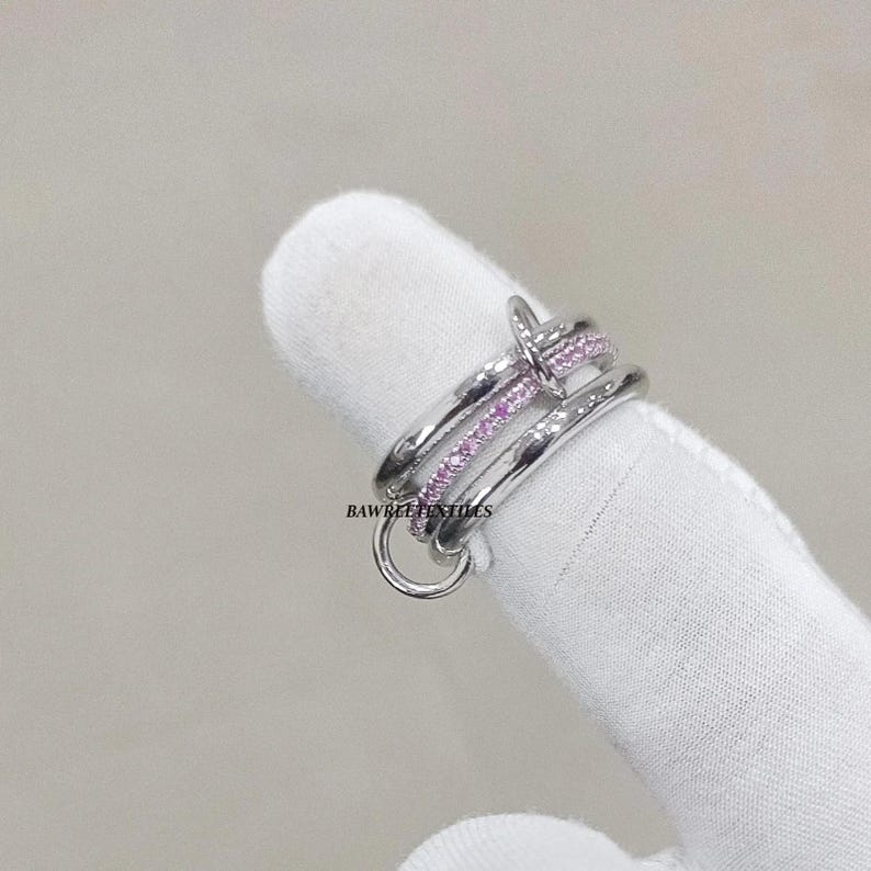 Pu&ograve; includere: Un anello color argento con un design a forma di chiodo. L'anello presenta tre fasce, una delle quali &egrave; adornata da piccole pietre preziose rosa. L'anello &egrave; mostrato su una superficie bianca.