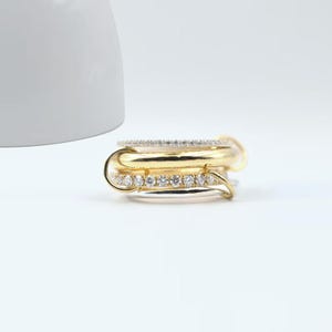 May include: A stack of gold and silver rings with clear gemstones. The rings are arranged in a layered design, with a mix of smooth and gemstone-studded bands. The rings are set against a white background.