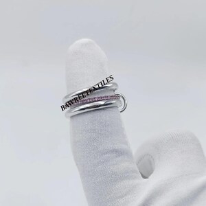 Pu&ograve; includere: Un anello d'argento con tre fasce, una delle quali incastonata con piccole pietre rosa. L'anello &egrave; su un dito coperto da un guanto bianco. Il testo "BAWREETEXTILES" &egrave; visibile nell'immagine.