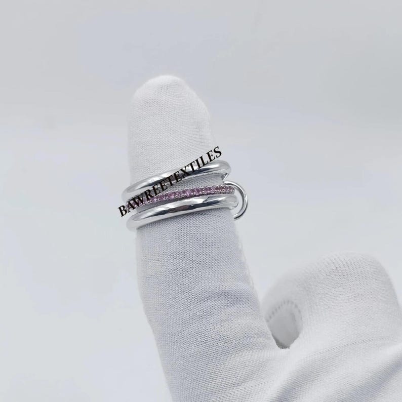 Pu&ograve; includere: Un anello d'argento con tre fasce, una delle quali incastonata con piccole pietre rosa. L'anello &egrave; su un dito coperto da un guanto bianco. Il testo "BAWREETEXTILES" &egrave; visibile nell'immagine.