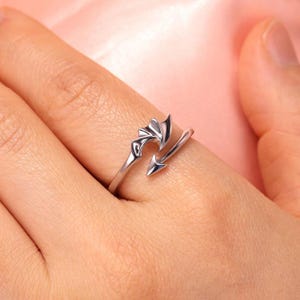 May include: A silver-toned open ring featuring a design with a pointed arrow tip on one end and a stylized wing or fin shape on the other. The metal band is polished and reflective, wrapping partially around the finger with a gap between the ends.
