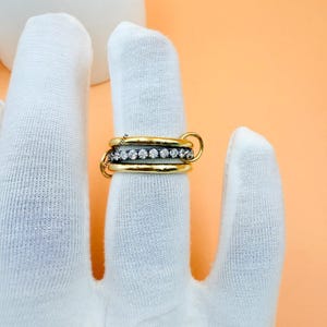 Op de afbeelding: Een goudkleurige ring met drie gestapelde banden. De middelste band heeft een rij kleine, heldere edelstenen. De ring wordt vastgehouden door een gehandschoende hand tegen een perzikkleurige achtergrond.