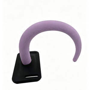 Puede incluir: Un gancho curvo de color morado claro con una textura sutil de veta de madera, unido a una placa de montaje rectangular negra. La placa cuenta con dos ranuras horizontales para su instalación, proporcionando una base para el gancho.