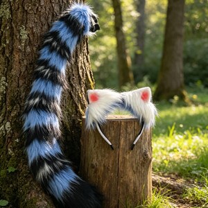 May include: A blue, black, and white faux fur tail leans against a tree trunk. A matching headband with white fur and pink inner ears sits on a wooden stump. The items are in a forest setting.