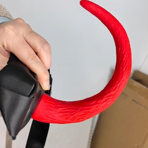 May include: A bright red, curved object with a textured surface, held by a black strap. The object's shape resembles a tail, with a rounded tip. The background is a plain white wall.