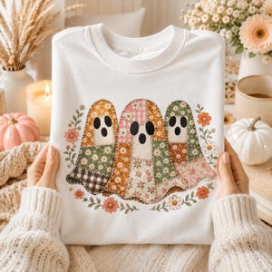 May include: A white crewneck sweatshirt featuring a graphic of three ghosts. Each ghost is composed of various patchwork fabric patterns including floral, gingham, and plaid prints in shades of orange, pink, green, and brown, surrounded by flowers.