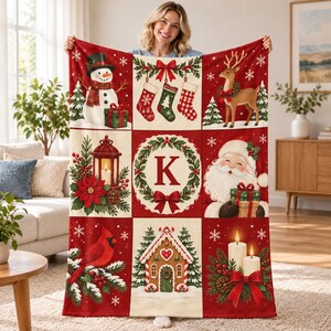 May include: A rectangular throw blanket featuring a grid of nine Christmas-themed illustrations including a snowman, stockings, a reindeer, a lantern, a wreath with the letter K, Santa Claus, a cardinal, a gingerbread house, and candles.