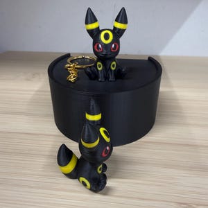 Può includere: Statuine di Umbreon nere con dettagli gialli e occhi rossi. Una è un portachiavi. Le statuine sono esposte su una piattaforma nera. Le figure di Umbreon sono su una superficie di legno marrone chiaro.
