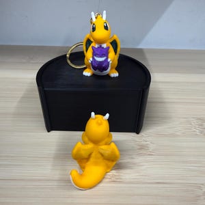 Può includere: Un portachiavi Dragonite giallo con una figura di Gengar viola e una statuetta di Dragonite gialla. Il portachiavi ha un anello dorato. Le statuette sono su una scatola nera e una superficie di legno.