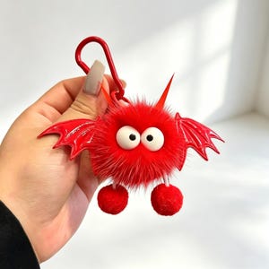 Puede incluir: Un llavero rojo y esponjoso con forma de monstruo de dibujos animados con ojos blancos grandes, alas rojas y dos pequeños pompones rojos. El llavero tiene un cierre de metal rojo.